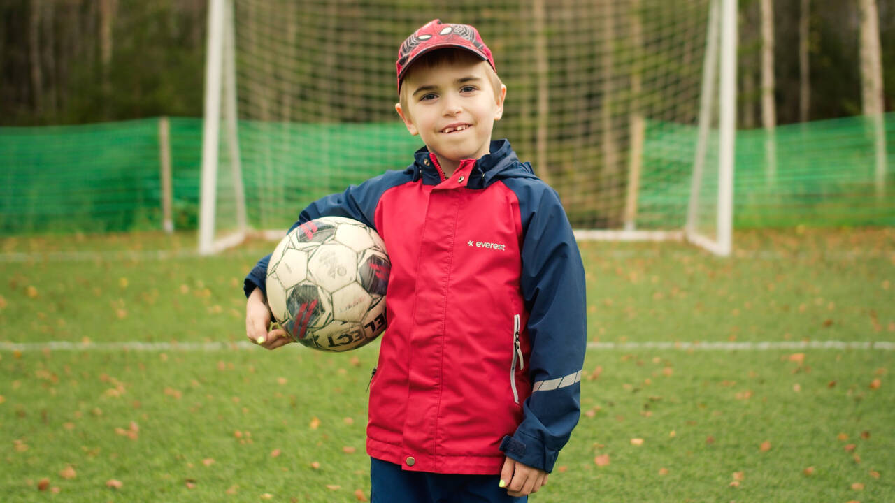 Poika pitä jalkapalloa kainalossa ja hymyilee kameralle.